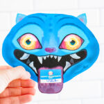 A hand is holding a single KPop Demon Hunters Inspired Valentine card shaped like a blue cartoon blue Derpy from KPop Demon Hunters. It's in front of a white tile surface.The Derpy looks like a blue tiger - has bright blue skin, pointed ears, glowing yellow-to-orange eyes, and a wide open mouth with sharp white teeth. A small Valentine card sticks out of its mouth like a tongue, with one reading &ldquo;Happy Valentine's Day."
