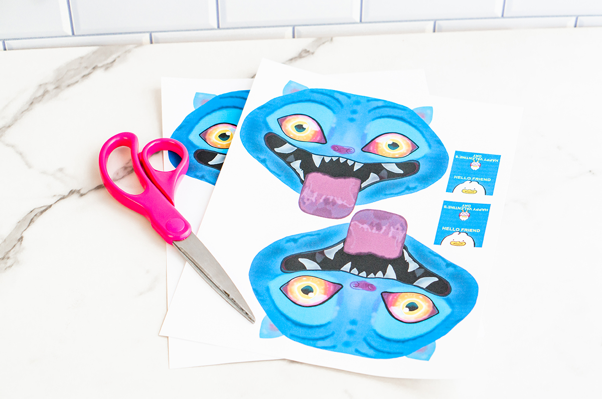 Two printed sheets of blue cartoon Derpy from KPop Demon Hunters Valentine designs lie slightly stacked on a white marble countertop with a white subway tile wall in the background. Each sheet shows a bright blue Derpy face with glowing yellow-orange eyes, pointed ears, and a wide open mouth with sharp white teeth and a purple tongue. Pink-handled scissors rest to the left, and small rectangular Valentine message cards are printed on the right side of the page, showing how the Valentine will be assembled.