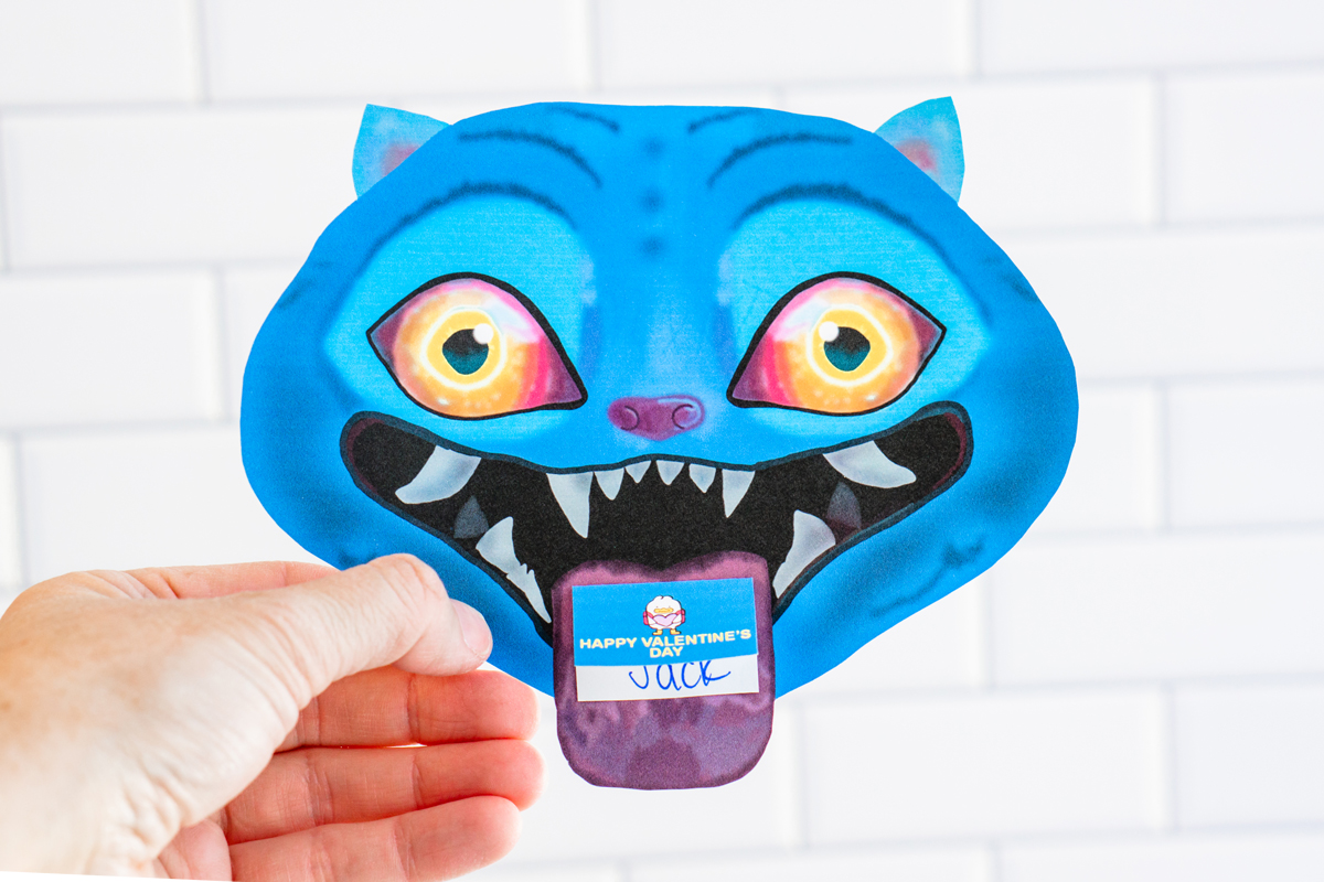 A hand is holding a single KPop Demon Hunters Inspired Valentine card shaped like a blue cartoon blue Derpy from KPop Demon Hunters. It's in front of a white tile surface.The Derpy looks like a blue tiger - has bright blue skin, pointed ears, glowing yellow-to-orange eyes, and a wide open mouth with sharp white teeth. A small Valentine card sticks out of its mouth like a tongue, with one reading &ldquo;Happy Valentine's Day."