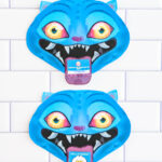 Two finished Valentine cards shaped like a blue cartoon blue Derpy from KPop Demon Hunters. They are placed on a white tile surface. Each demon has bright blue skin, pointed ears, glowing yellow-to-orange eyes, and a wide open mouth with sharp white teeth. A small Valentine card sticks out of each demon&rsquo;s mouth like a tongue, with one reading &ldquo;Happy Valentine&rsquo;s Day&rdquo; and the other reading &ldquo;Hello Friend."