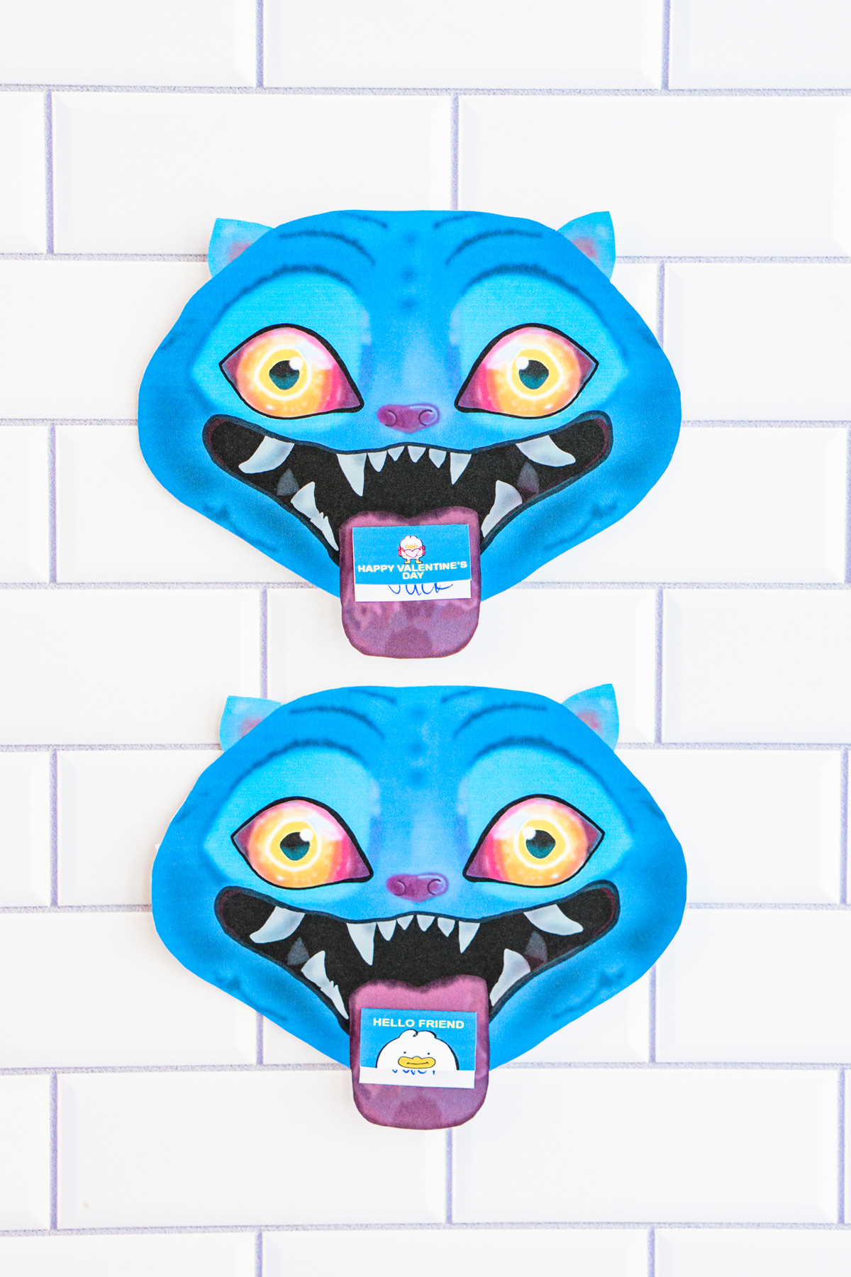 Two finished Valentine cards shaped like a blue cartoon blue Derpy from KPop Demon Hunters. They are placed on a white tile surface. Each demon has bright blue skin, pointed ears, glowing yellow-to-orange eyes, and a wide open mouth with sharp white teeth. A small Valentine card sticks out of each demon&rsquo;s mouth like a tongue, with one reading &ldquo;Happy Valentine&rsquo;s Day&rdquo; and the other reading &ldquo;Hello Friend."