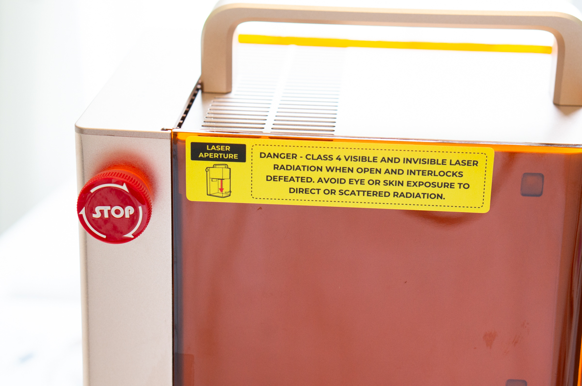 Front corner of the xTool F2 showing the emergency stop button and laser safety warning label.