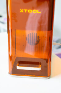 Front view of the xTool F2 powered on with the illuminated xTool logo visible through the orange safety enclosure.
