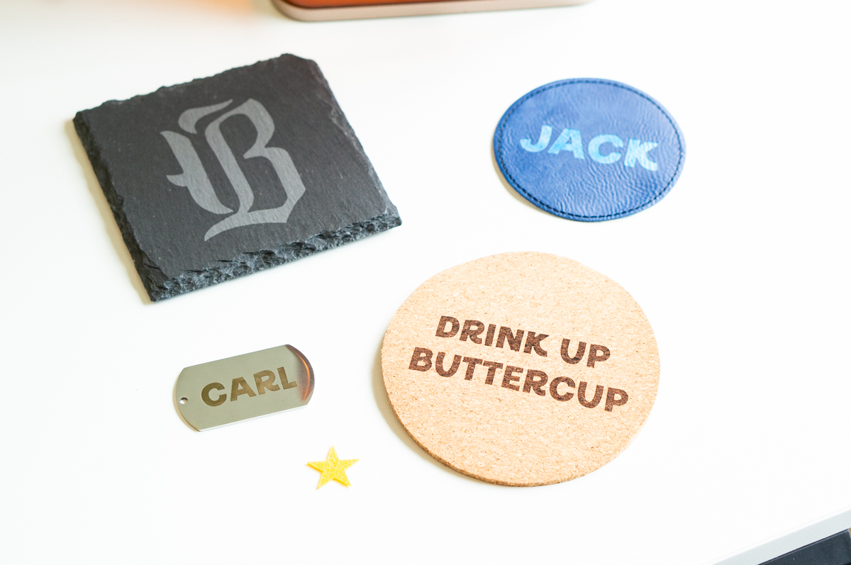 Close-up of engraved xTool F2 sample projects including a slate coaster, leather coaster, cork coaster, and metal dog tag.
