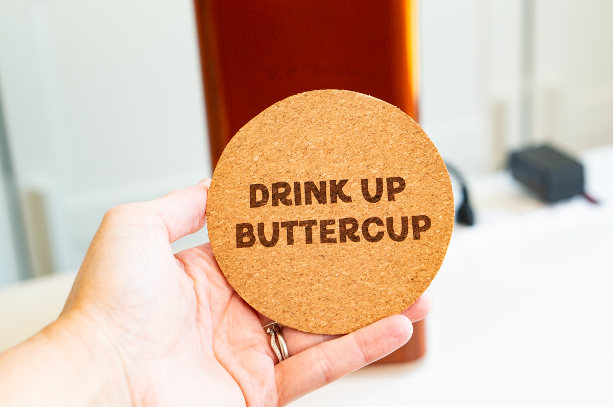 Hand holding a cork coaster engraved with the phrase "Drink Up Buttercup" using the xTool F2 laser engraver.