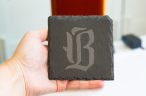 Hand holding a slate coaster engraved with a decorative monogram using the xTool F2 laser engraver.