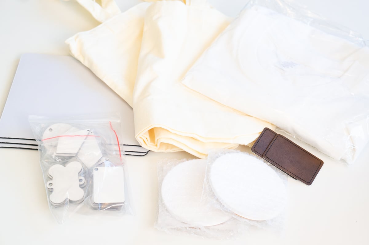 Assortment of blank craft items spread across a white table, including cream and white fabric tote bags, white fabric items, square coasters, round coasters, rectangular tags, and small metal shapes. The unfinished blanks can be personalized with vinyl, sublimation, laser engraving, or other crafting techniques.