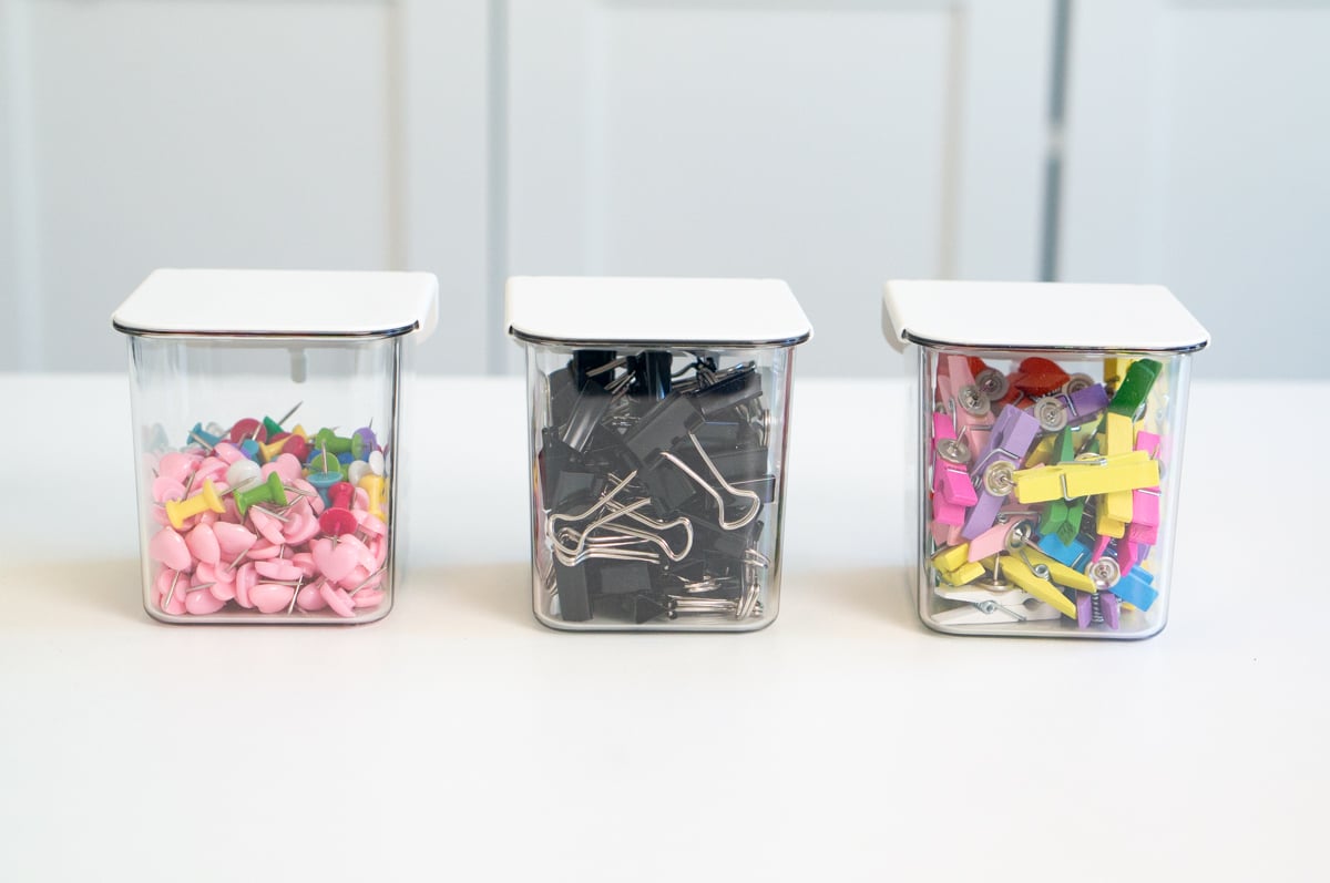 Three clear square storage containers with white lids arranged on a white table. The left container is filled with colorful push pins, including many pink heart-shaped pins, the center container holds black binder clips in various sizes, and the right container contains small wooden clothespins painted in bright colors including pink, yellow, purple, green, and blue.