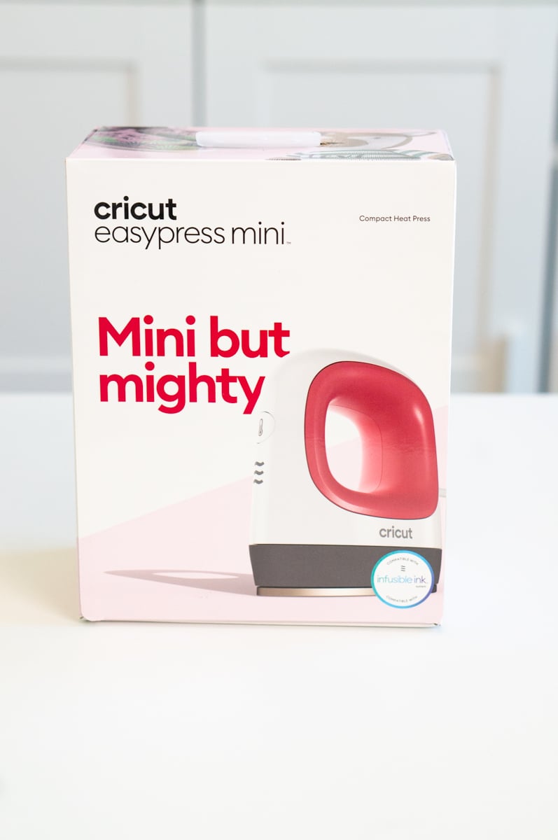 Box for a Cricut EasyPress Mini compact heat press displayed upright on a white table. The white and pale pink packaging shows the small handheld heat press with a red handle and includes the phrase