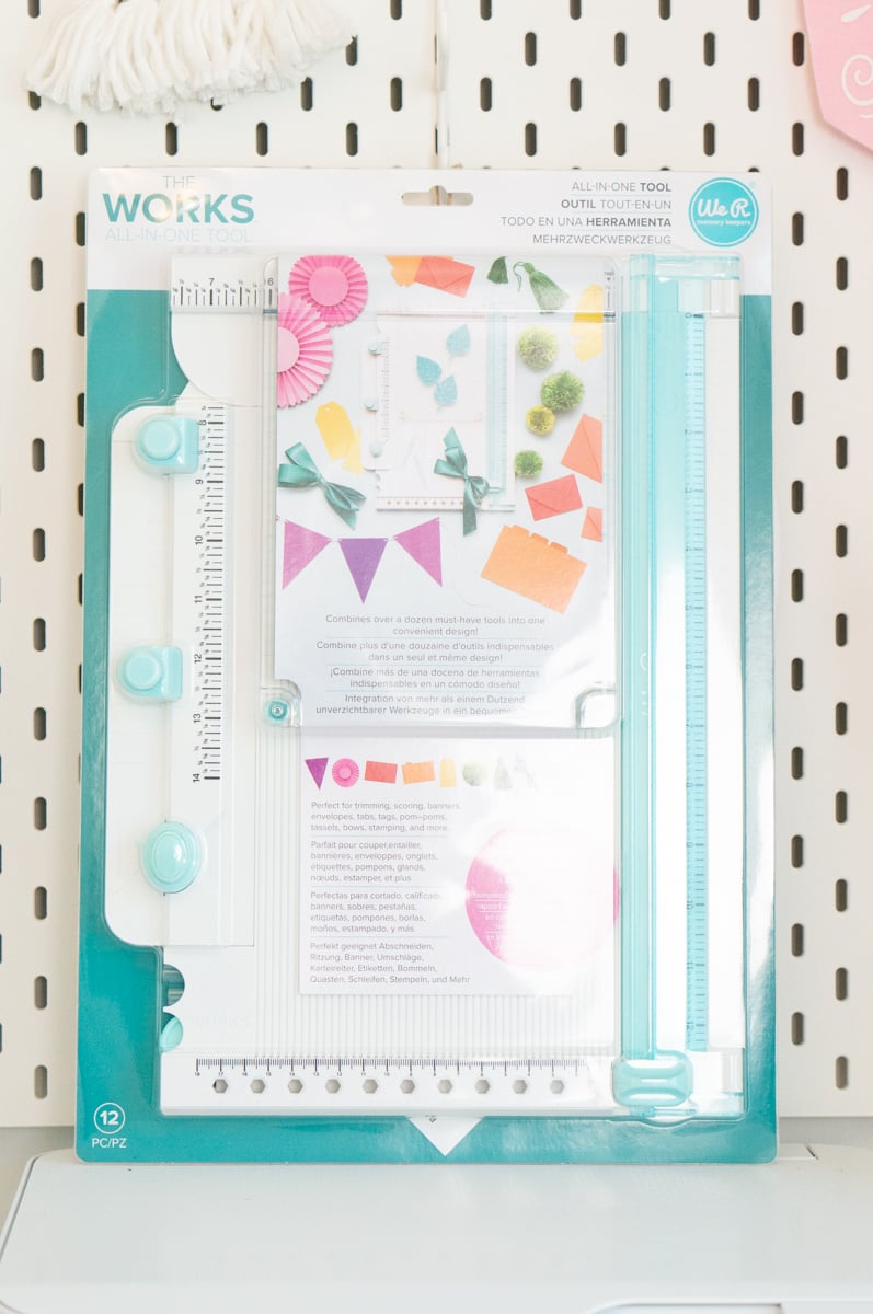 We R Memory Keepers The Works All-in-One Tool displayed in its original turquoise and white packaging against a white pegboard. The multifunction paper crafting tool includes rulers, guides, a cutting and scoring area, and other features for creating envelopes, tags, bags, pom-poms, tassels, bows, banners, and additional paper crafts.