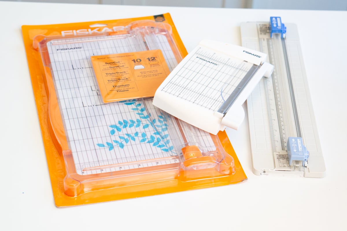 Assortment of three Fiskars paper cutters arranged on a white table. The collection includes a large orange and clear rotary-style paper trimmer in its packaging, a smaller white tabletop paper cutter with a measurement grid, and a long narrow paper trimmer with a clear ruler and blue sliding cutting blade.