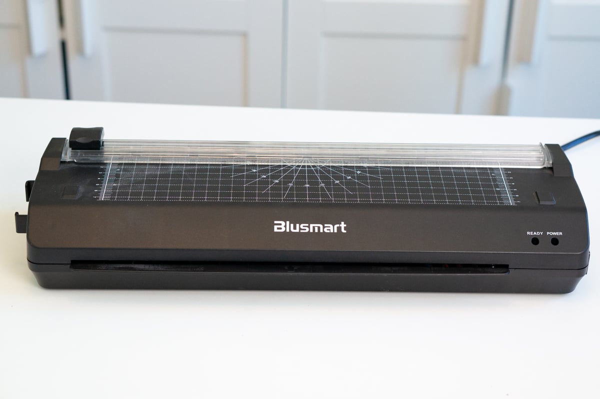 Black Blusmart laminating machine sitting on a white table in front of pale blue cabinets. The wide desktop laminator has a measurement grid and angle guides across the top, a clear paper guide, power and ready indicator lights, and a front opening where laminated pages exit.