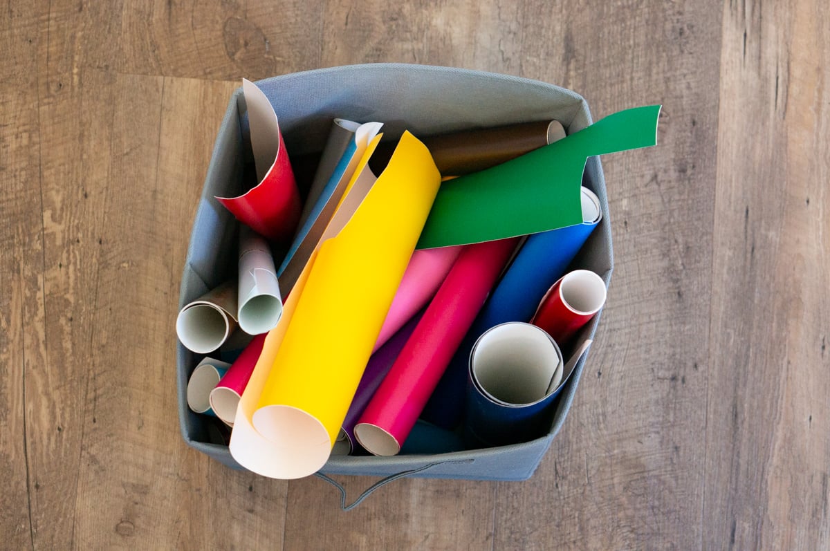 Gray fabric storage bin filled with partially used rolls and scraps of colorful craft vinyl. The rolls include bright yellow, pink, red, green, blue, black, white, and other colors and are stored upright and at different angles inside the bin on a wood floor.