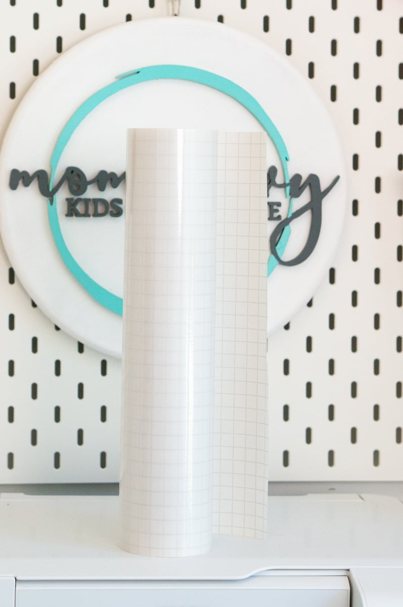 Large roll of clear gridded transfer tape standing upright on a white shelf in front of a white craft room pegboard. The transparent transfer tape has a faint square grid to help with positioning and transferring adhesive vinyl designs onto craft projects.