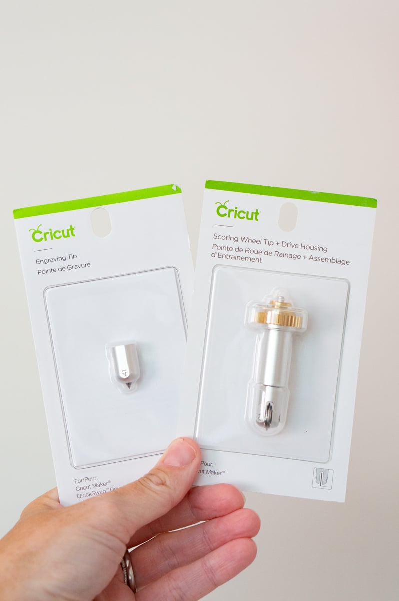 A hand holding two unopened Cricut specialty machine tools against a plain light background. One package contains a Cricut Engraving Tip, and the other contains a Cricut Scoring Wheel Tip with Drive Housing, showing examples of specialty blades and tools that can expand the types of projects made with a compatible Cricut machine.