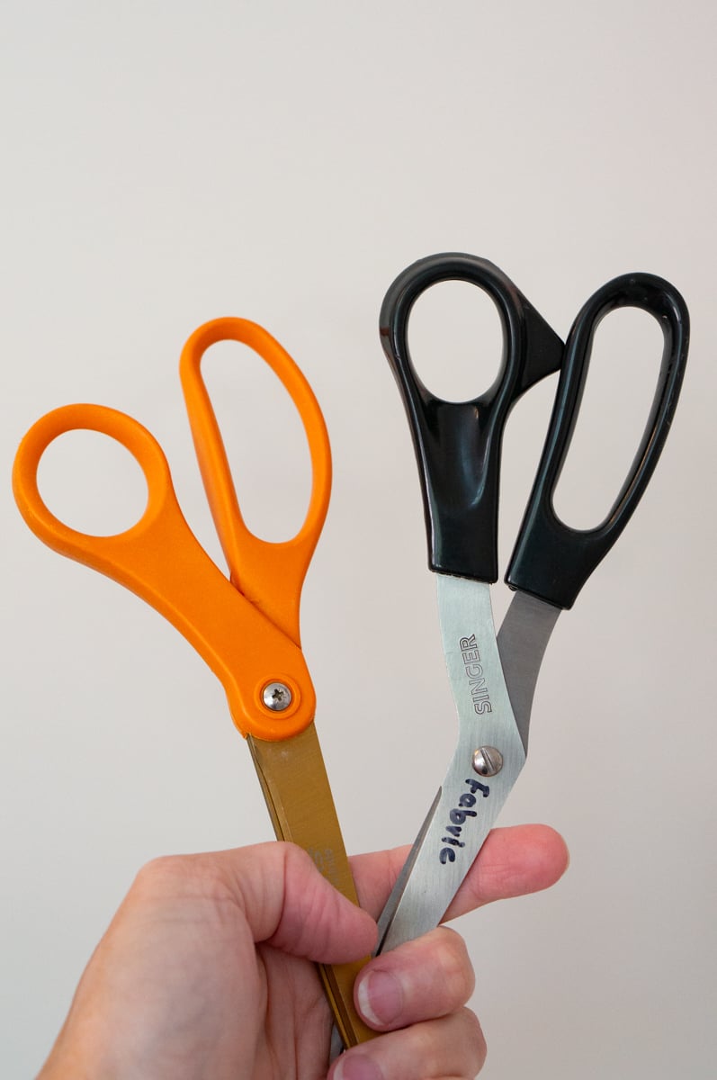 A hand holding three pairs of craft scissors against a plain light background. The scissors include a large pair with bright orange handles and two pairs with black handles, including a pair of Singer fabric scissors, showing different sizes and styles of scissors used for general crafting, fabric cutting, and other projects.