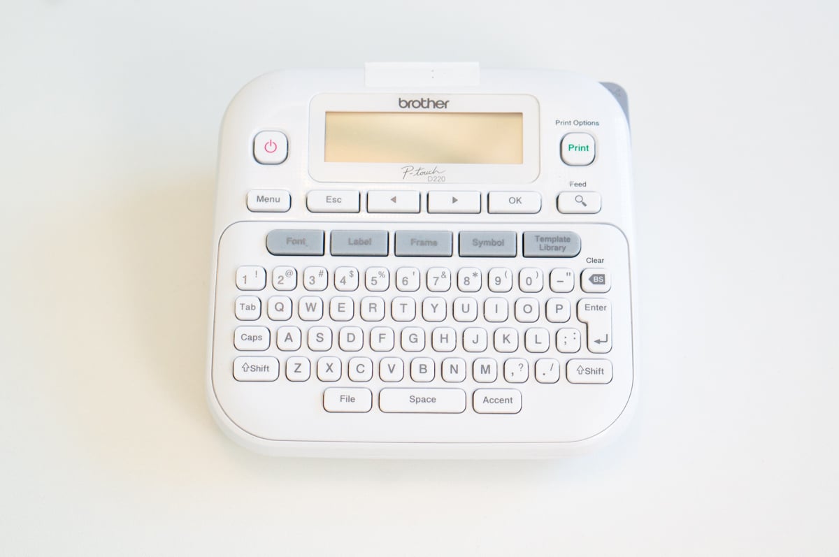 White Brother P-touch D220 label maker photographed from above on a white surface. The compact label maker has a built-in QWERTY keyboard, display screen, print button, and buttons for selecting fonts, labels, frames, symbols, and templates. A small printed label reading