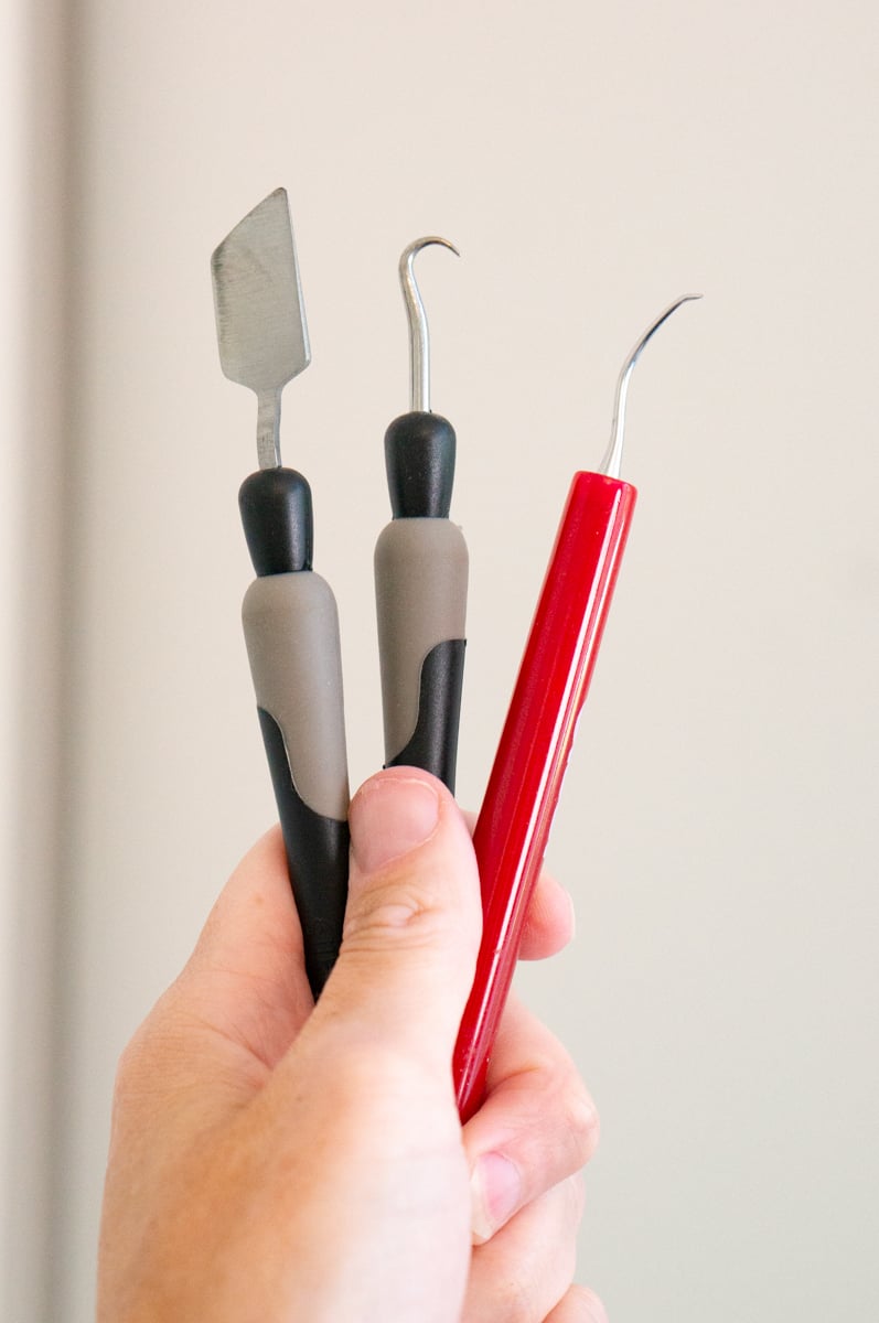 A hand holding three vinyl crafting and weeding tools against a plain light background. The tools include a wide metal scraper or spatula with a gray and black handle, a hooked weeding tool with a gray and black handle, and a fine curved metal pick with a red handle for removing small pieces of vinyl and working with detailed designs.