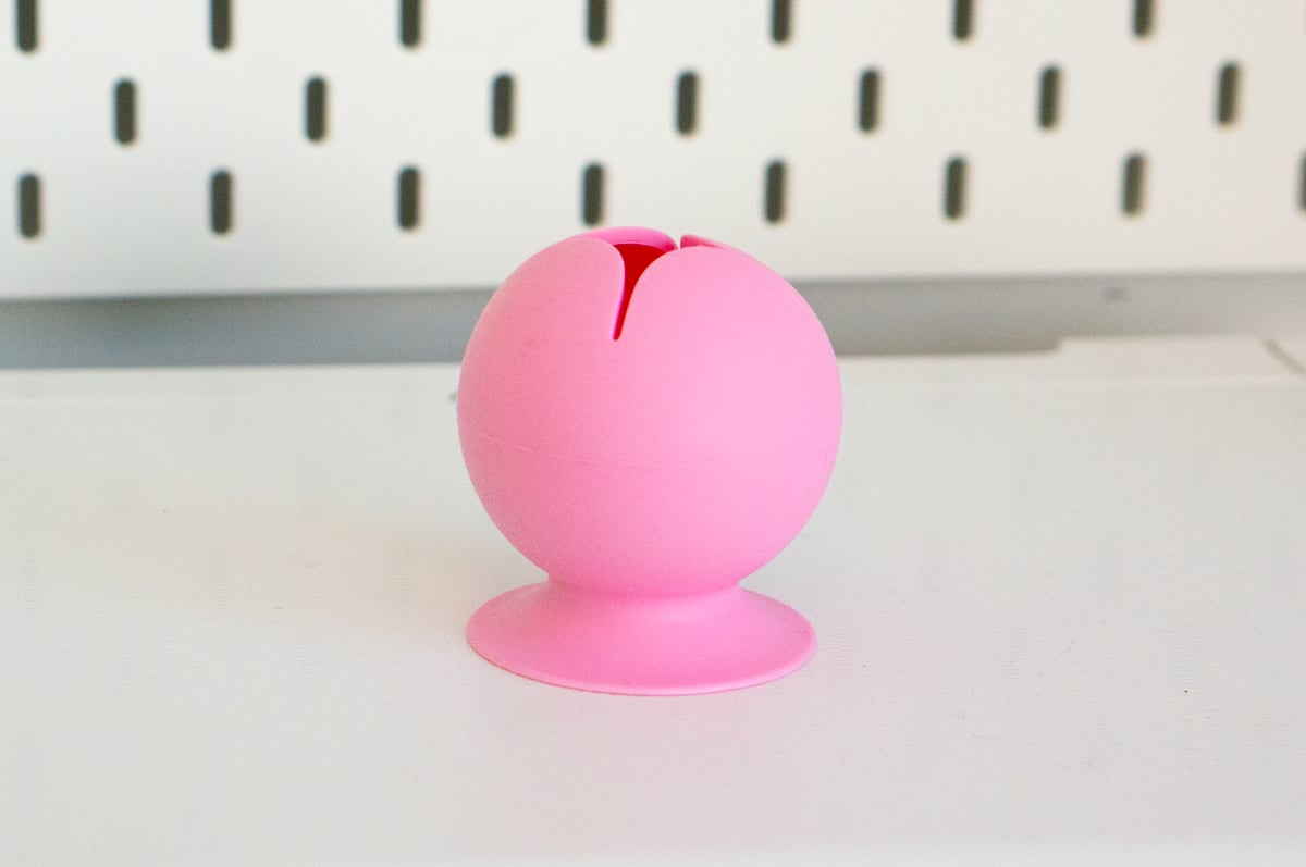 Small bright pink silicone vinyl scrap collector sitting on a white craft table in front of a white pegboard. The round cup has a narrow opening at the top for scraping off small pieces of vinyl while weeding and a wide suction-cup base that holds it in place on the work surface.