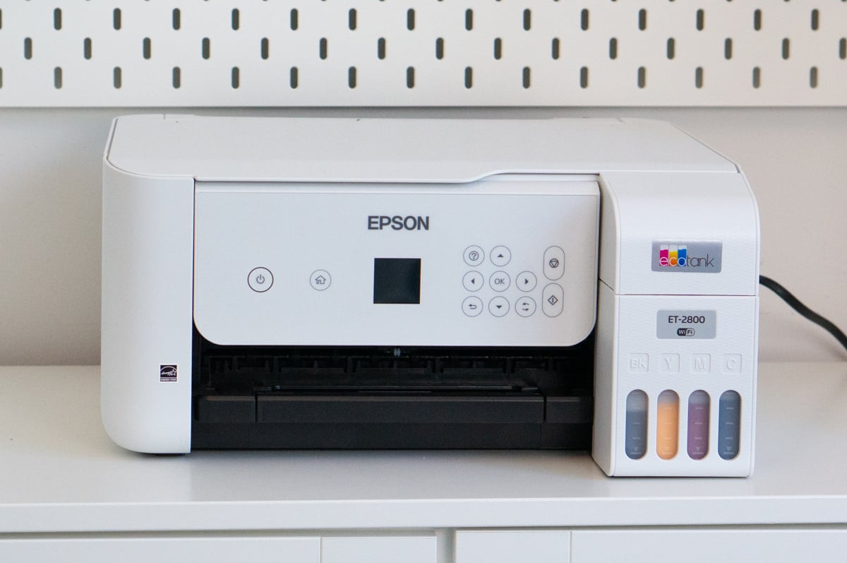 White Epson EcoTank ET-2800 color printer sitting on a white shelf in front of a white pegboard. The printer has a small control screen and buttons on the front, an open paper output area, and four visible refillable ink tanks for black, yellow, magenta, and cyan ink on the right side.