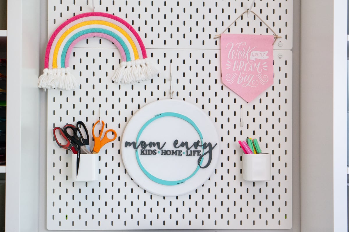Large white craft room pegboard organized with crafting tools and colorful decorations. The pegboard holds several pairs of scissors in a white cup, colorful pens and markers in another cup, a handmade rainbow with pink, yellow, white, turquoise, and lavender arches and white fringe, a round Mom Envy sign, and a pink hanging banner that reads