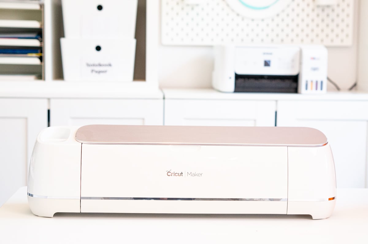 White Cricut Maker craft cutting machine with a rose gold lid sitting on a white craft table. A white Epson printer, labeled storage bins, and a white pegboard are visible in the organized craft room behind the machine.