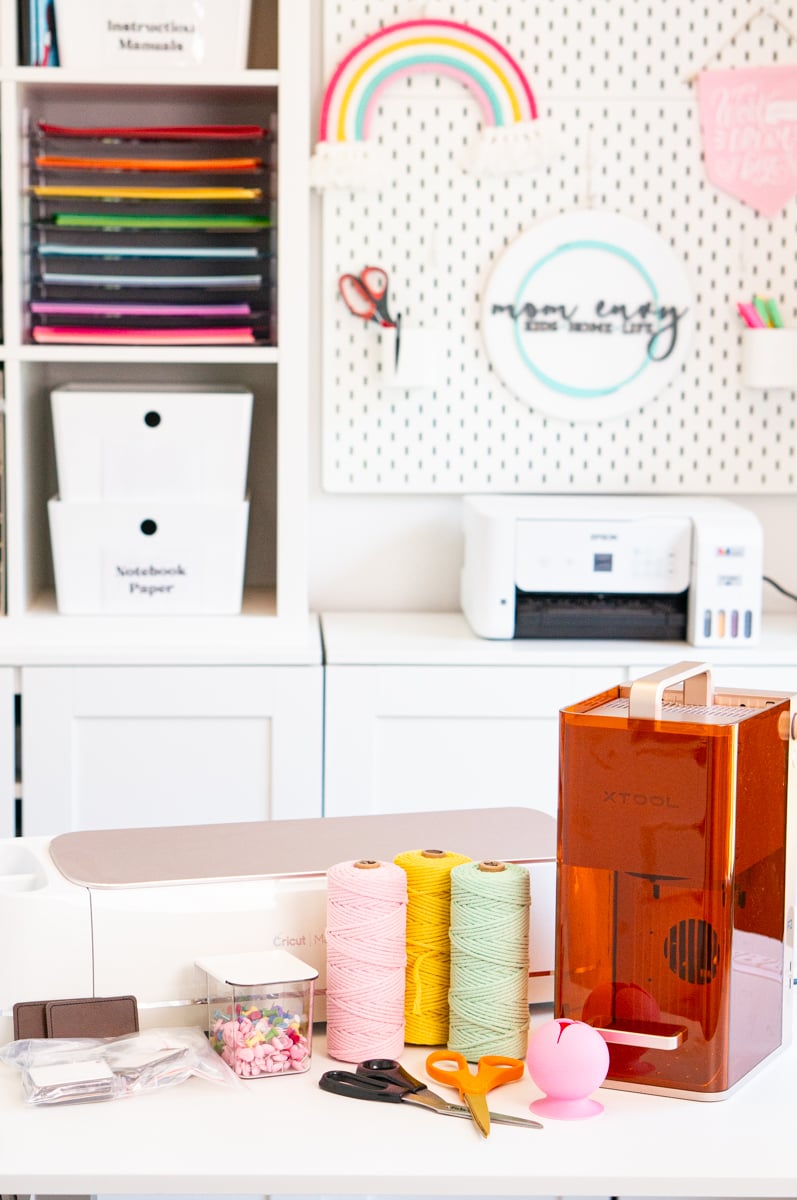 Assortment of gift ideas for crafters displayed on a white craft table in a bright, organized craft room. Items include a white Cricut Maker cutting machine, an orange-covered xTool portable laser machine, pink, yellow, and mint green spools of craft twine, black and orange craft scissors, a pink vinyl scrap collector, a clear storage container filled with colorful push pins, and several blank craft items ready to personalize. An Epson printer, craft paper storage, and decorated pegboard are visible in the background.