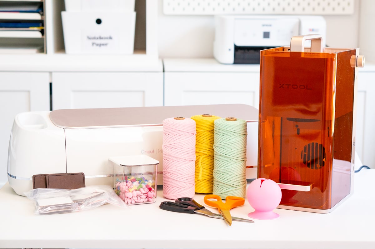Close-up display of craft gift ideas arranged on a white table, including a white Cricut Maker cutting machine, orange-covered xTool portable laser machine, three large spools of pink, yellow, and mint green craft twine, black and orange scissors, a pink silicone vinyl scrap collector, clear storage container filled with colorful push pins, and several blank items for crafting and personalization. An organized white craft room with storage bins and a printer is softly visible in the background.
