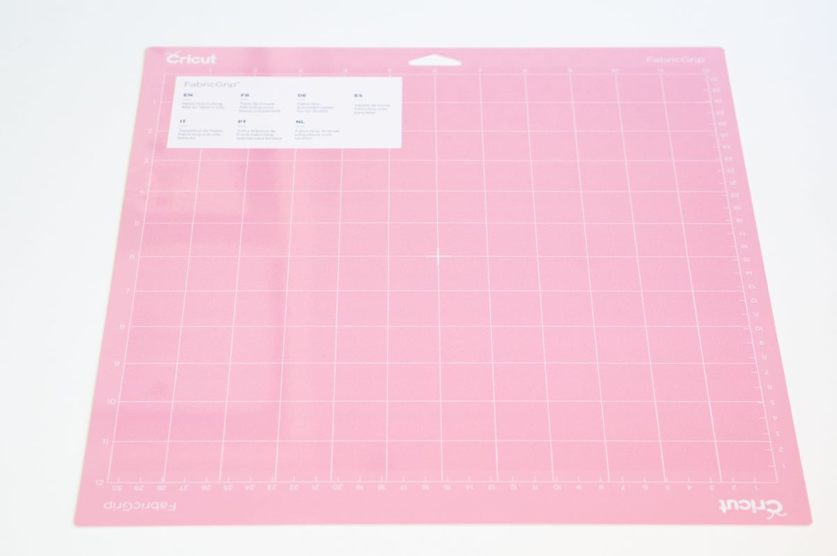 Pink Cricut FabricGrip cutting mat lying on a white surface. The rectangular craft machine mat has a white measurement grid with inch markings around the edges and is designed to hold fabric in place while cutting with a compatible Cricut machine.