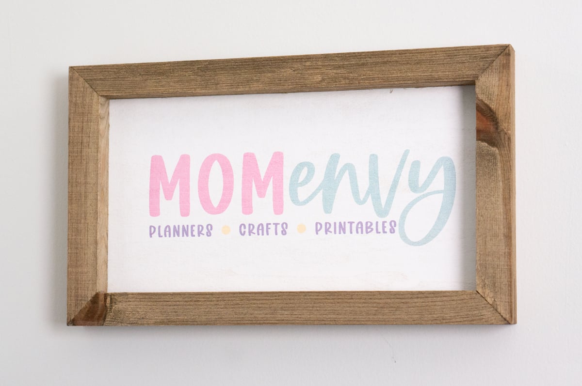 Custom rectangular Mom Envy craft room sign hanging on a white wall. The handmade sign has a rustic stained wood frame surrounding a white background with the colorful Mom Envy logo in pink and light blue and the words