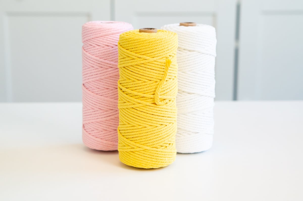 Three large spools of colorful cotton craft twine displayed on a white table in front of pale blue cabinets. The spools are pink, bright yellow, and white and represent basic craft supplies that can be given as gifts, such as twine, ribbon, yarn, and other crafting materials.