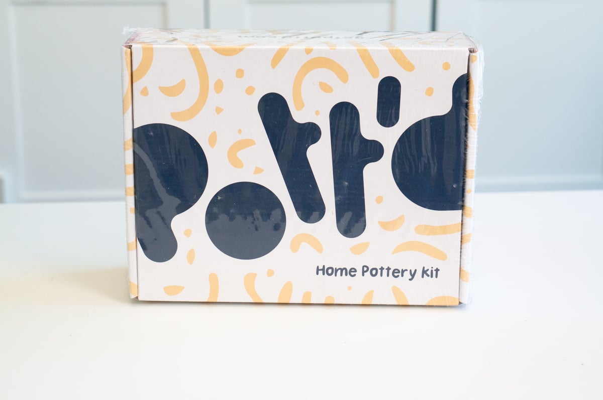 Boxed Pott'd Home Pottery Kit sitting on a white table in front of pale blue cabinets. The pre-made DIY clay craft kit comes packaged in a cream-colored box decorated with abstract yellow and dark blue shapes and is an example of a craft kit that can be given to someone who enjoys trying new creative projects.