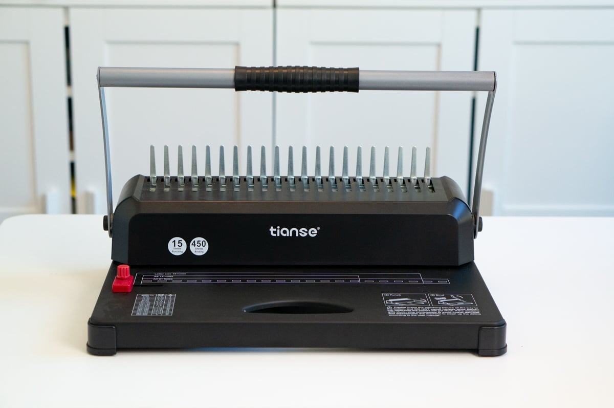 Black Tianse manual binding machine sitting on a white table. The desktop machine has a long metal handle, a row of binding punches, and guides for punching holes into paper and assembling handmade notebooks, planners, journals, and other bound paper projects.