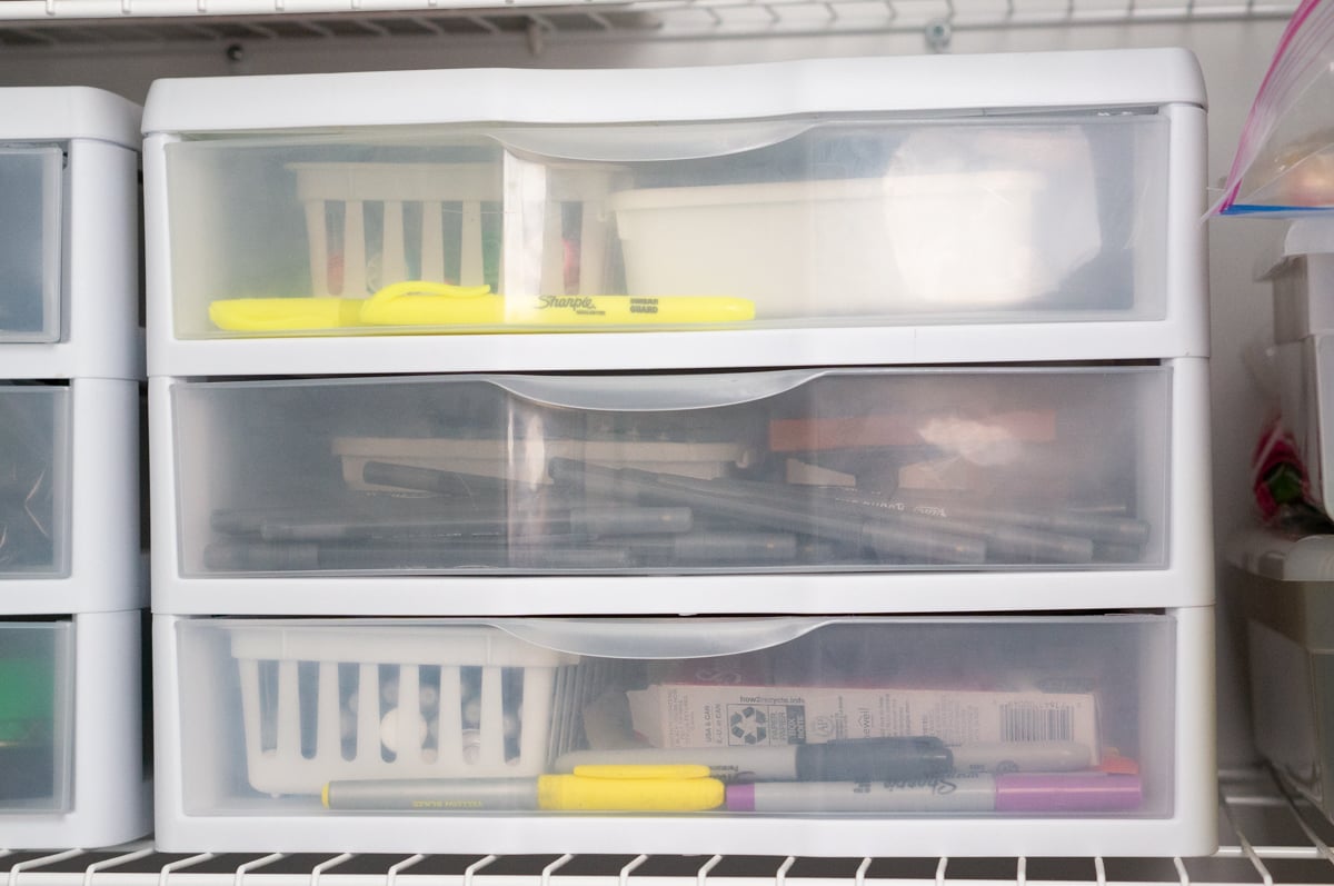 White plastic three-drawer craft storage unit filled with pens, markers, tools, adhesives, and other crafting supplies. The translucent drawers make it possible to see the supplies stored inside while keeping small craft materials organized.