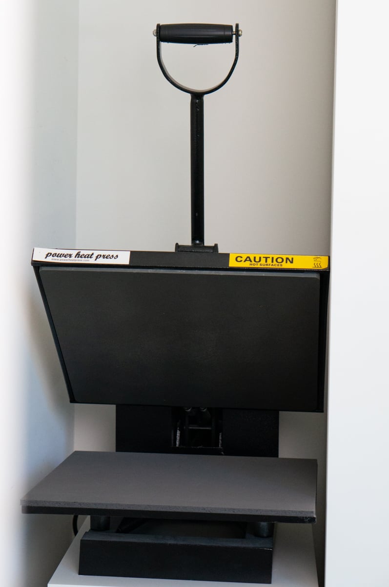 Large black Power Heat Press machine stored in a craft room cubby. The full-size clamshell-style heat press has a large upper heating plate, padded lower platen, and long handle and can be used to apply heat transfer vinyl and other heat-applied designs to shirts, tote bags, and craft blanks.