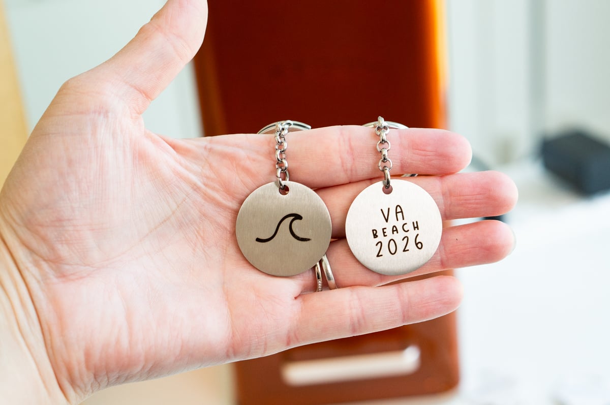 Two custom laser engraved metal keychains featuring a wave design and VA Beach 2026 text