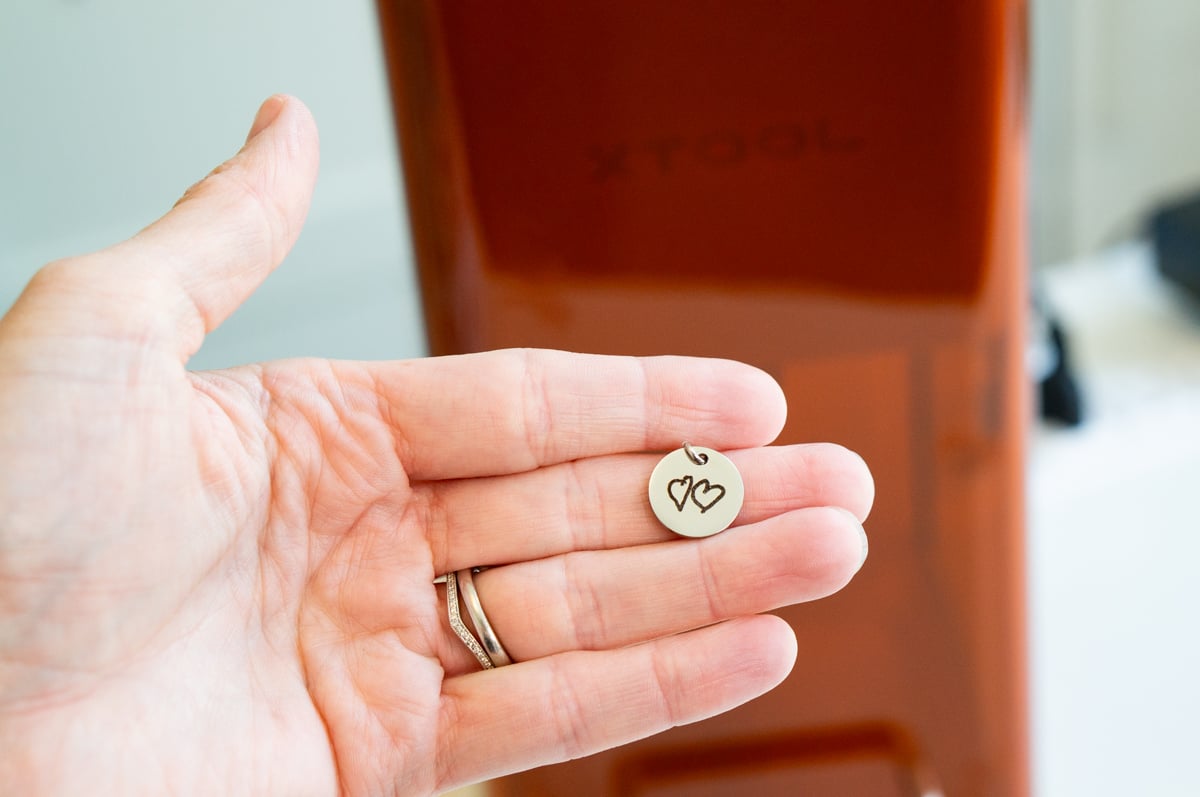 This is an image of a round, silver pendant being held in a hand that has been engraved using the xTool F2. The engraving are two hearts hand drawn by children. 
