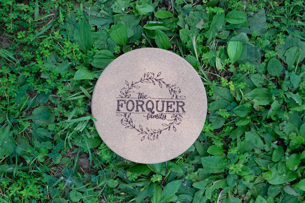 Round cork trivet engraved with The Forquer Family inside a leafy wreath, photographed outdoors surrounded by green plants.