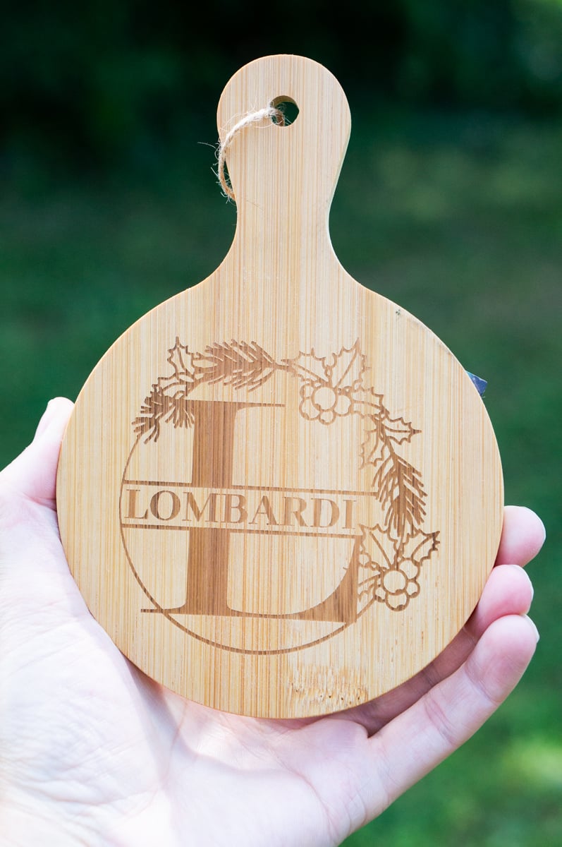 Hand holding a small bamboo charcuterie board engraved with a large letter L, the family name Lombardi, and a Christmas wreath of pine branches and holly berries.