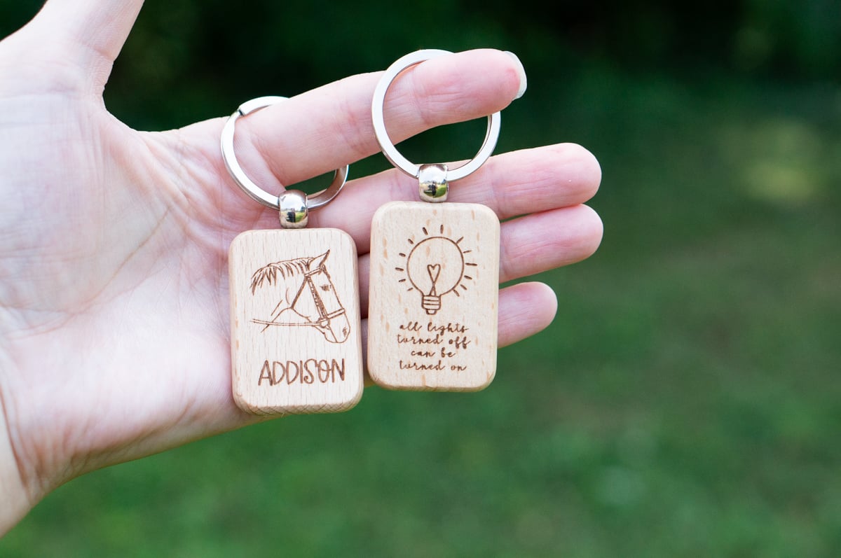Hand holding two personalized wooden keychains, one engraved with a horse and the name Addison and the other with a light bulb and the phrase all lights turned off can be turned on.