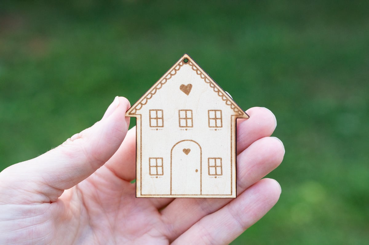 Hand holding a laser cut wooden house ornament engraved with windows, a rounded front door, small hearts, and decorative scalloped trim along the roof.