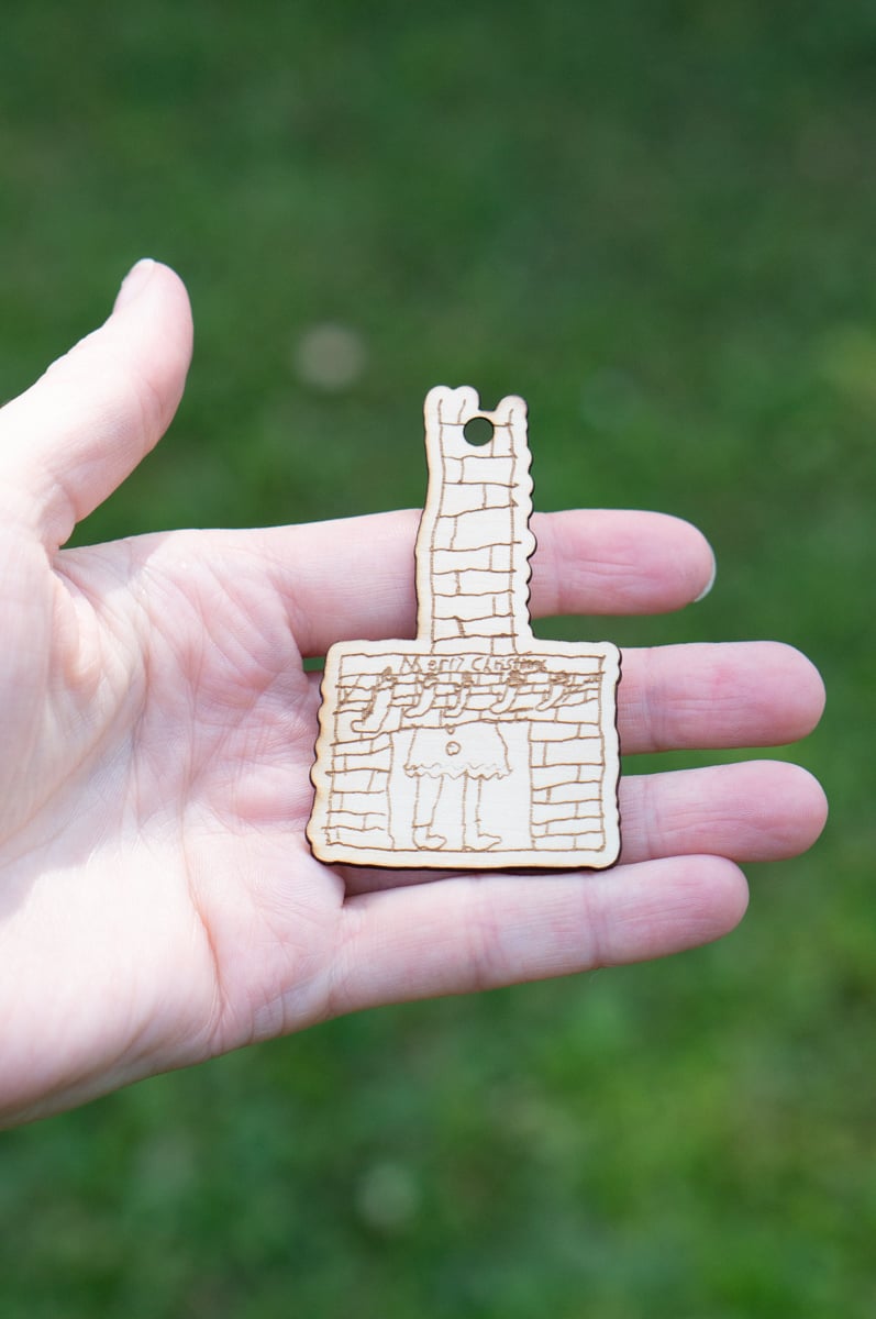 Hand holding a laser cut wooden ornament made from a child's hand-drawn picture of a brick fireplace and tall chimney.