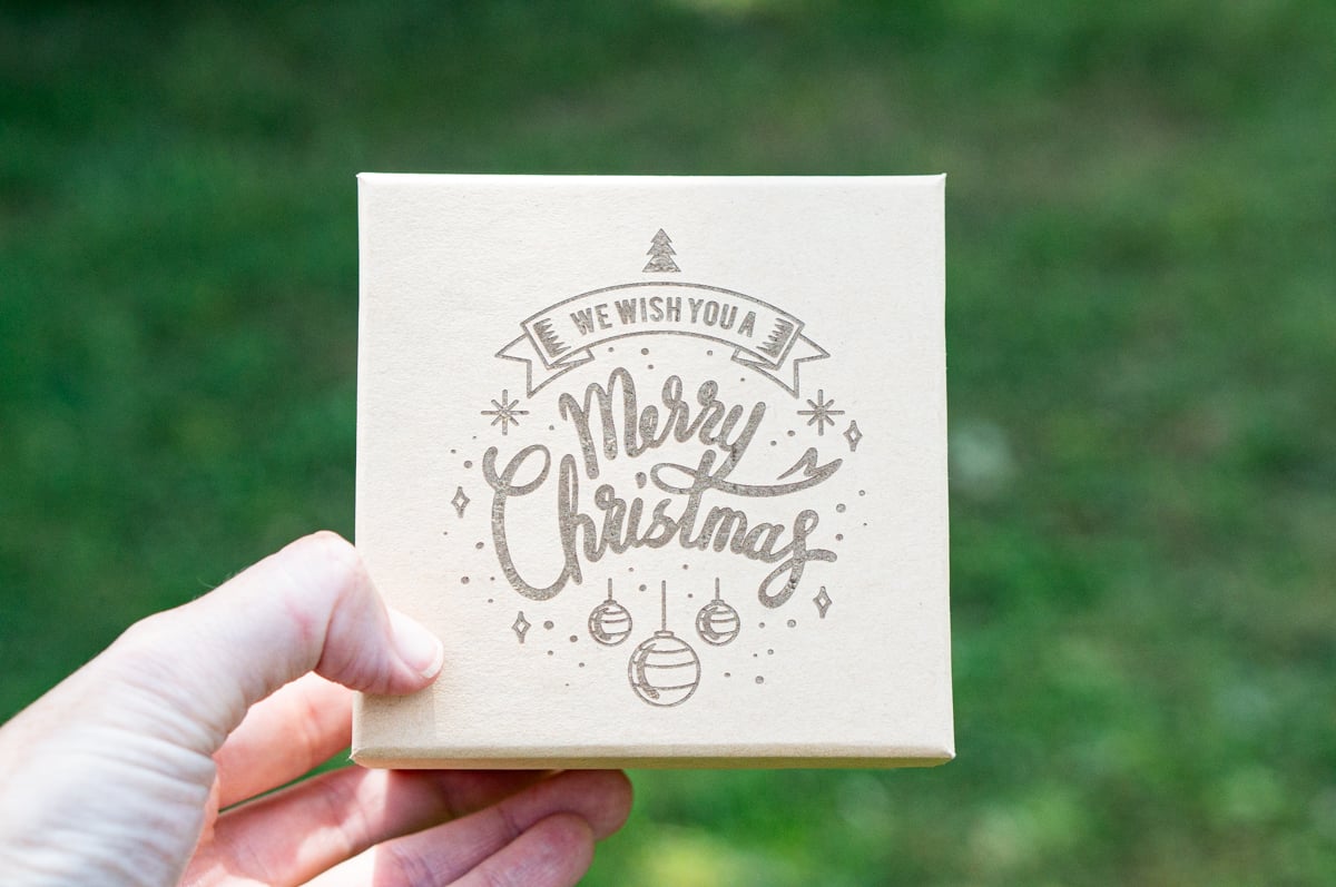 Hand holding a kraft gift box laser engraved with We Wish You a Merry Christmas, surrounded by Christmas ornaments, snowflakes, and a small Christmas tree.