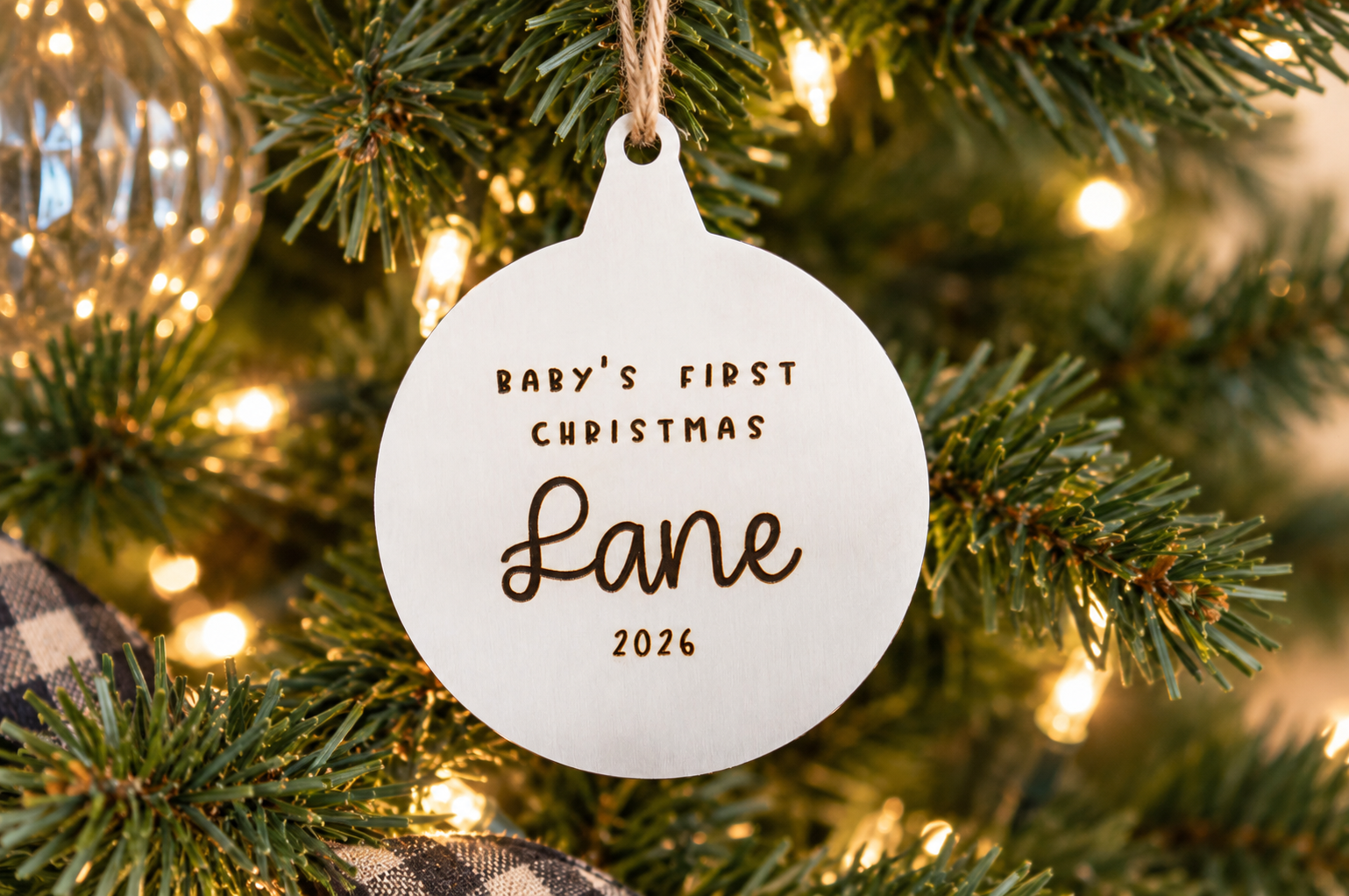 Personalized silver metal Baby's First Christmas ornament engraved with the name Lane and the year 2026, hanging from a lit Christmas tree.