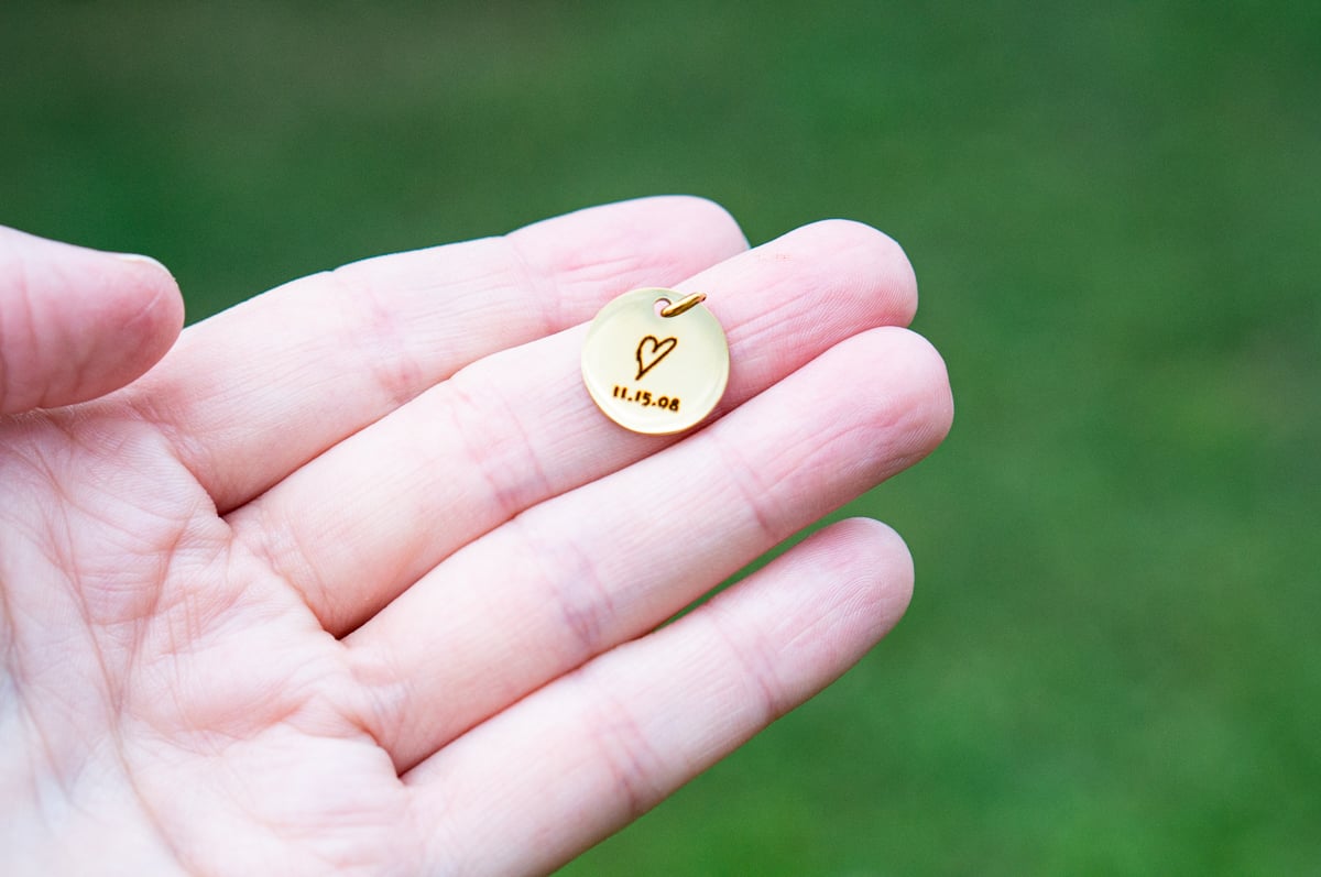 Hand holding a small round gold necklace charm engraved with a hand-drawn heart and the date 11.15.08 against a blurred green background.