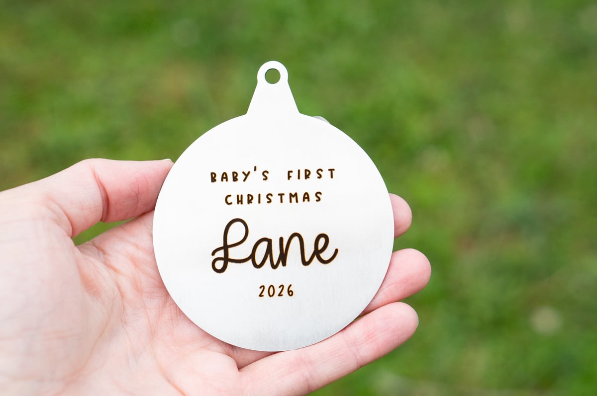 Hand holding a large round silver metal Christmas ornament engraved with Baby's First Christmas, the name Lane, and the year 2026.
